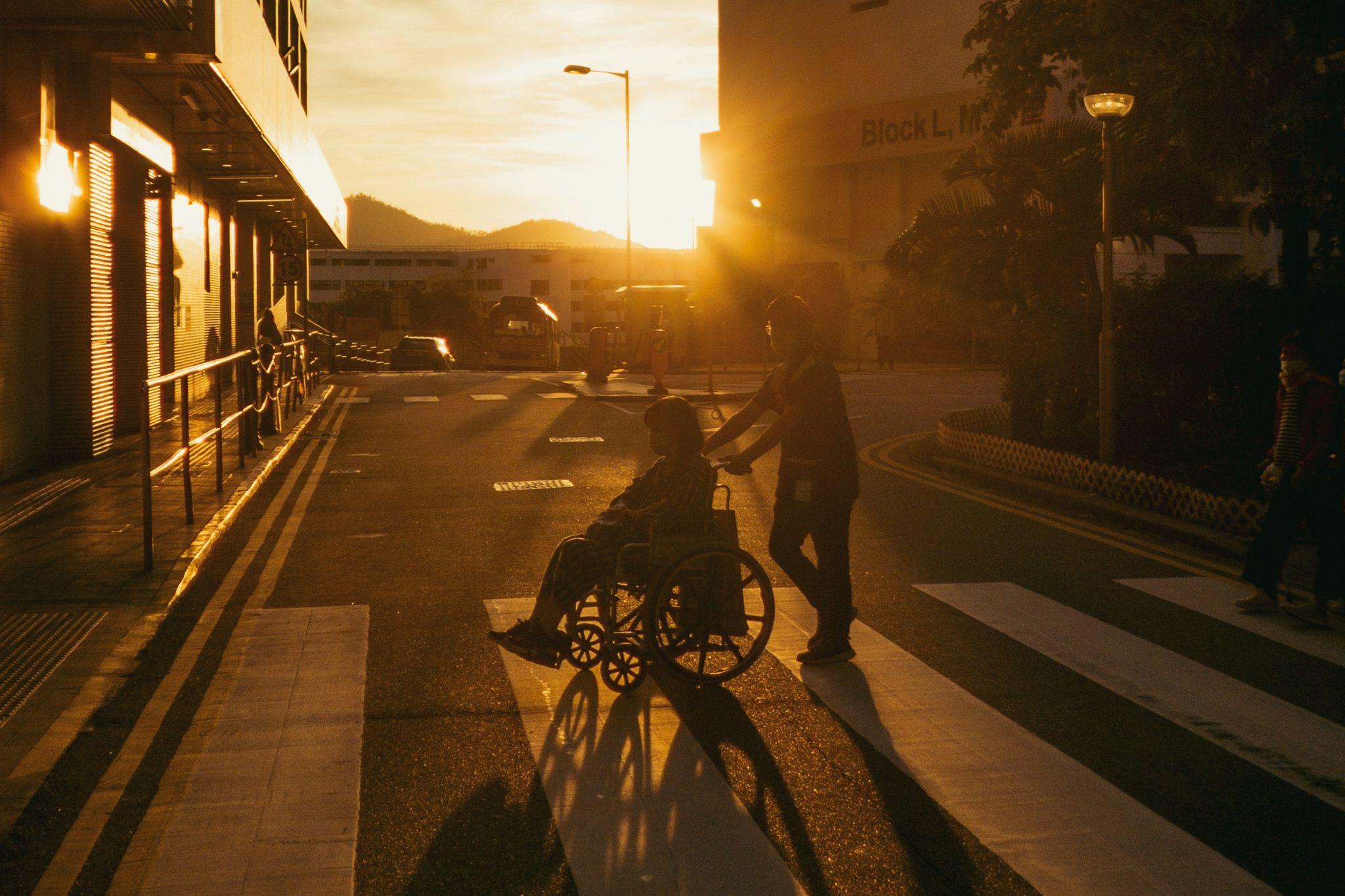 A person sits in a wheelchair, pushed by another person across a zebra crossing. The sun is low in the sky so they're almost silhouettes on the city street 