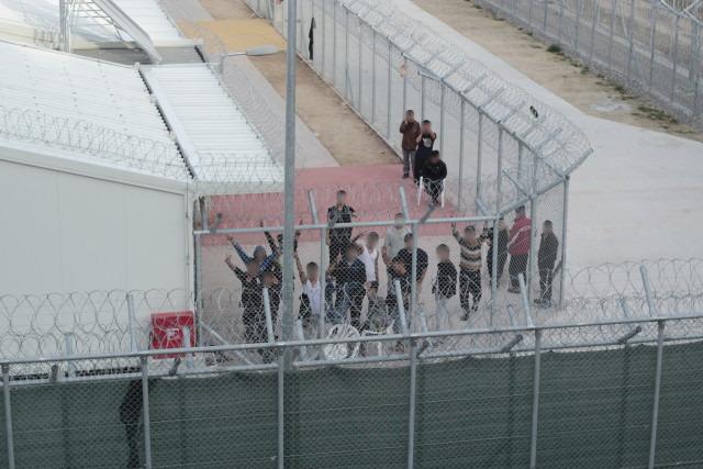 People can be seen standing in a small yard with two layers of barbed-wire-topped fences keeping them in. They wave their arms and turn their faces (blurred) to the sky.