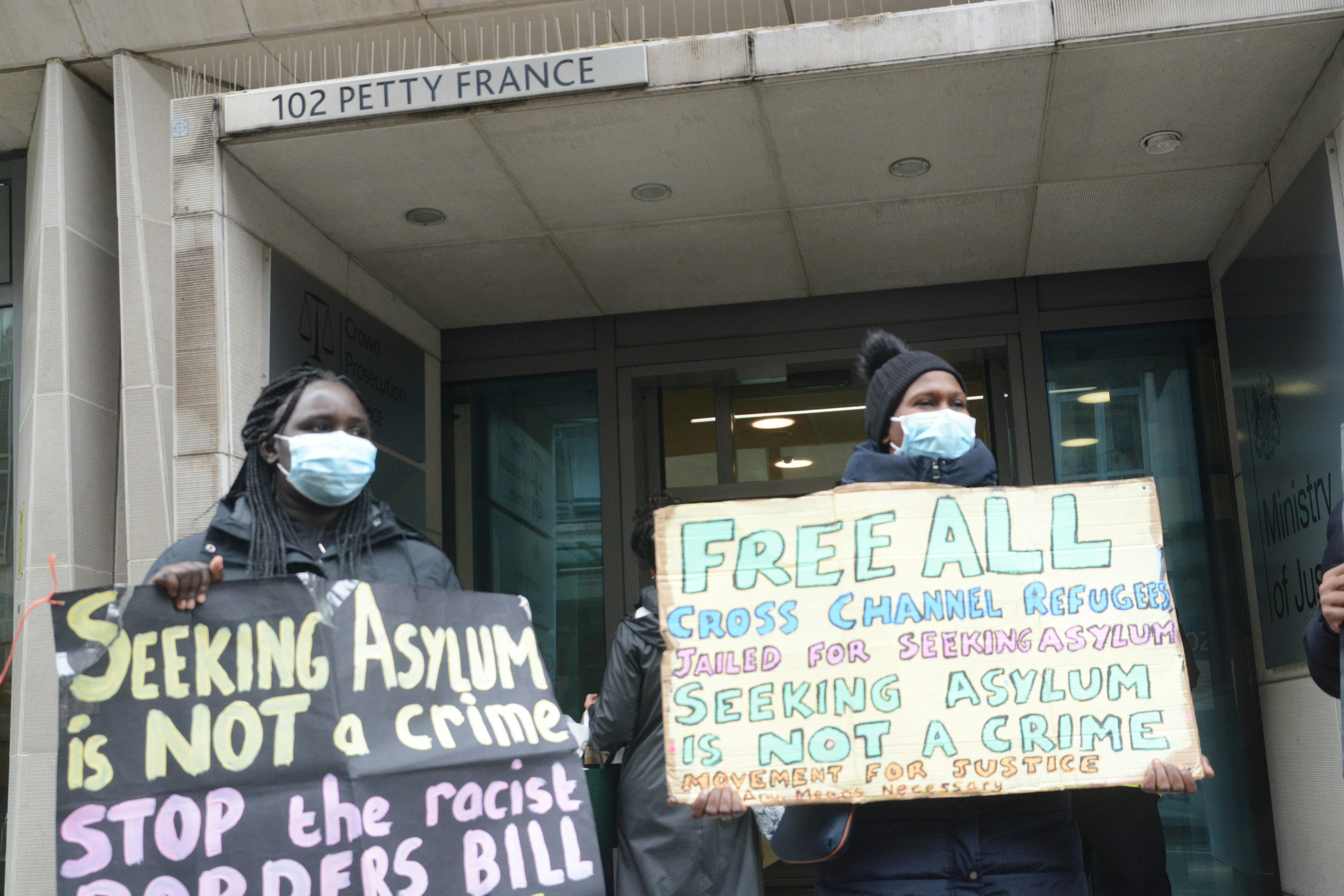 Two people wearing thick coats and masks hold up signs reading: seeking asylum is not a crime. stop the racist borders bill. Free all cross channel refugees jailed for seeking asPhoto by <a href="https://unsplash.com/@piprobins?utm_source=unsplash&utm_medium=referral&utm_content=creditCopyText">Philip Robins</a> on <a href="https://unsplash.com/photos/a-group-of-people-standing-outside-of-a-building-holding-signs-TjSgpqhk74I?utm_source=unsplash&utm_medium=referral&utm_content=creditCopyText">Unsplash</a>   