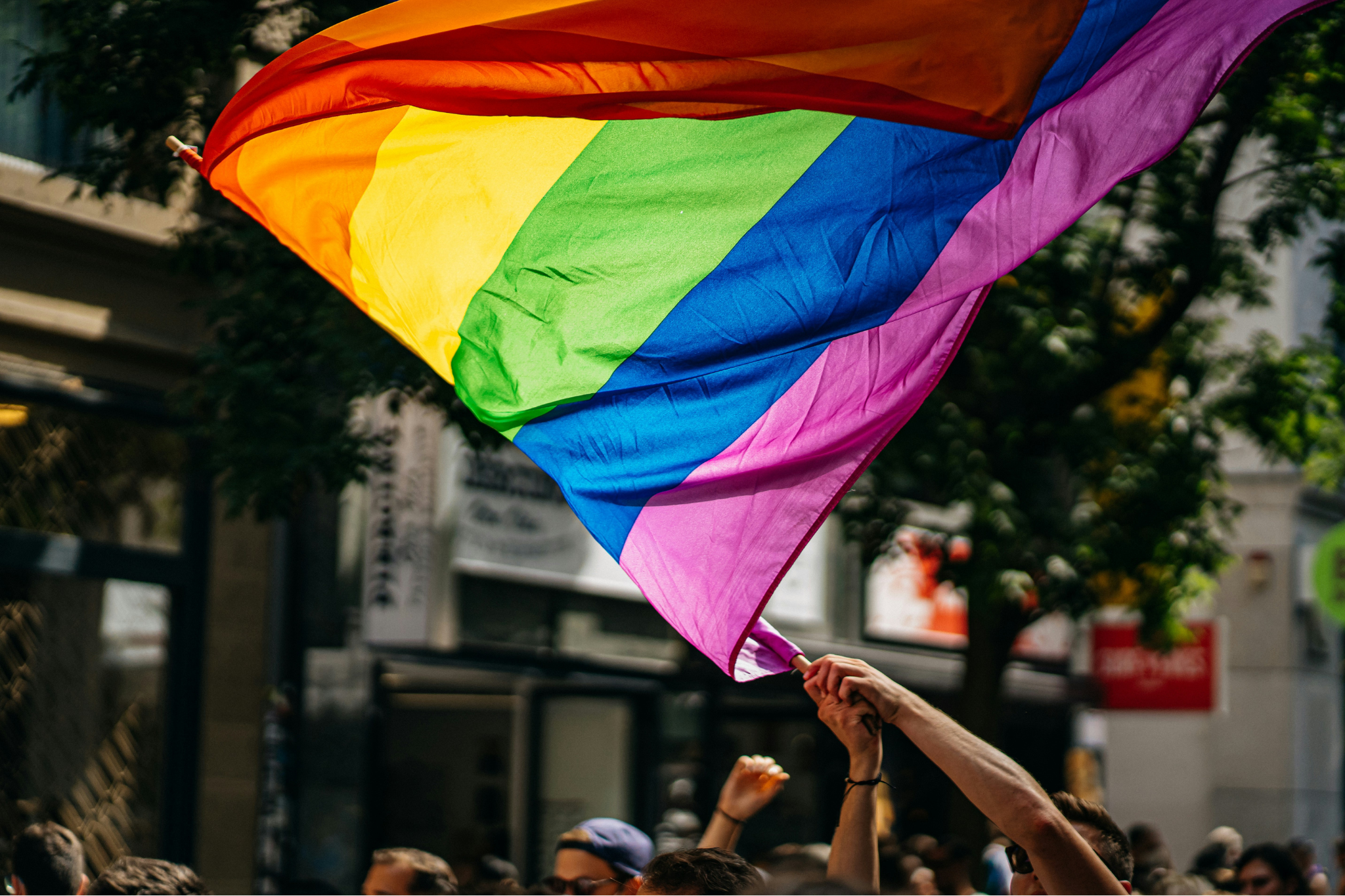 A person's arm above a crowd of people, waving a rainbow LGBTQ+ Pride flag in a city street