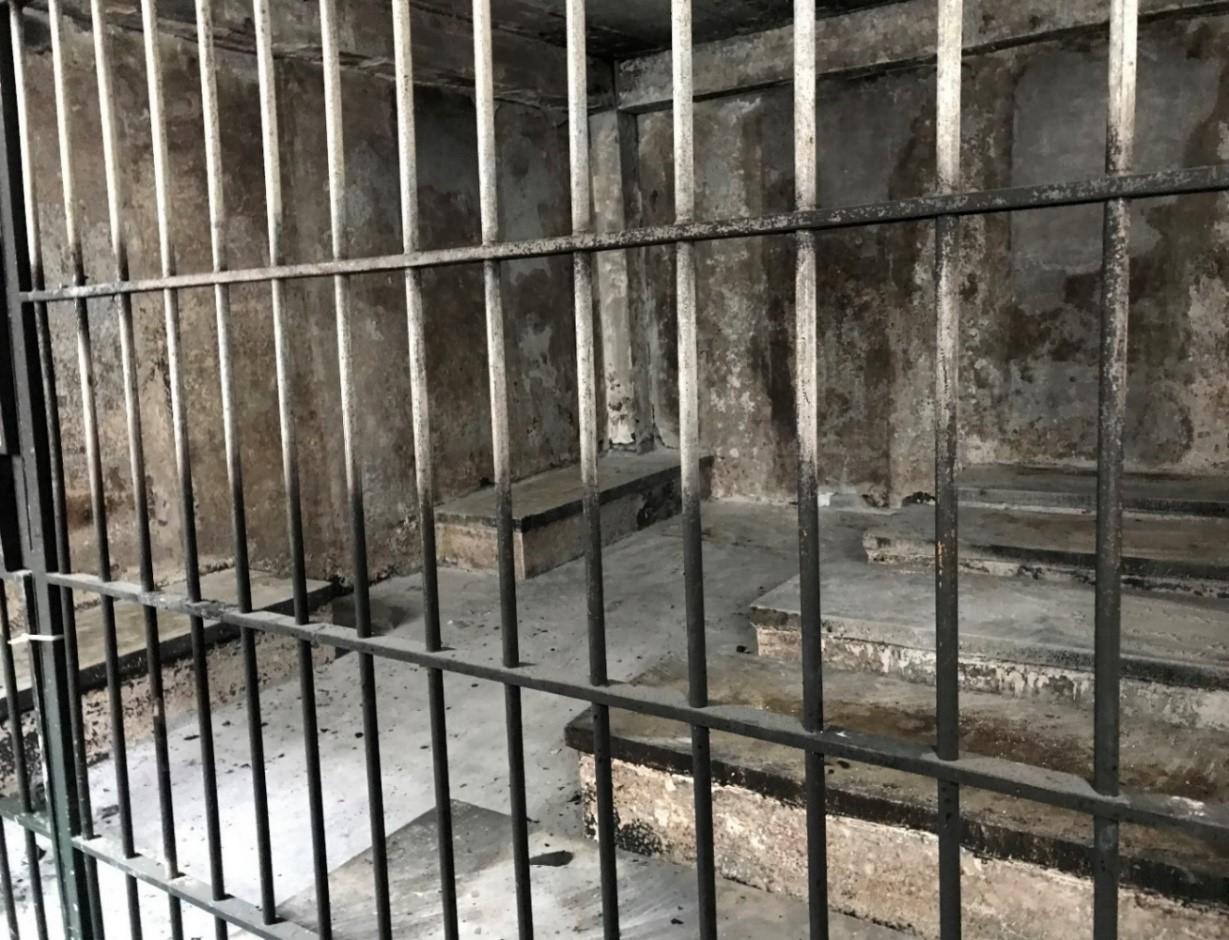 Inside of what looks like an abandoned cell, as seen through the bars. Conditions look terrible, there are no people or furnishings inside. Everything is dirty