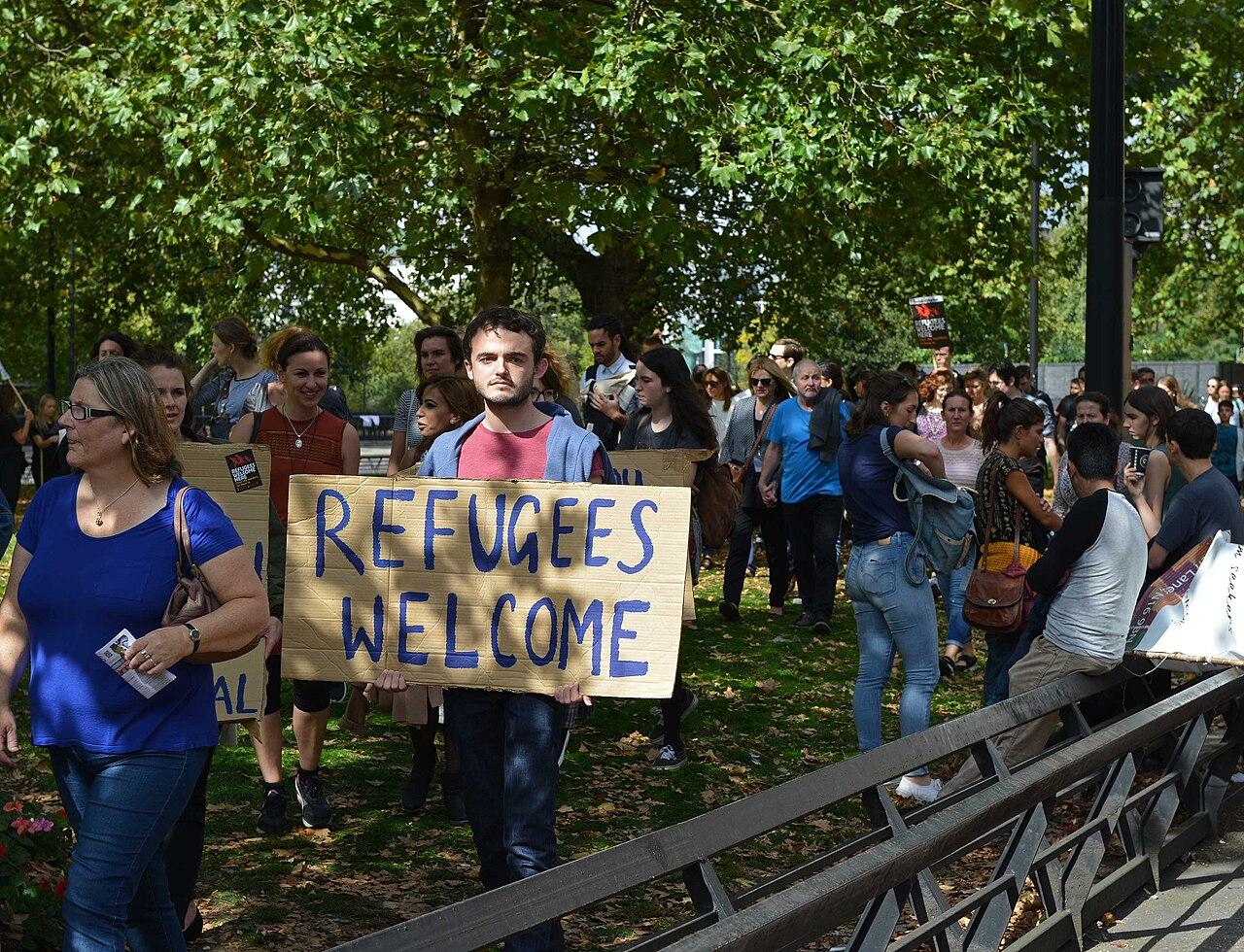 A man holds a cardboard sign reading Refugees Welcome. He stands under trees with other people around, some also holding cardboard placards