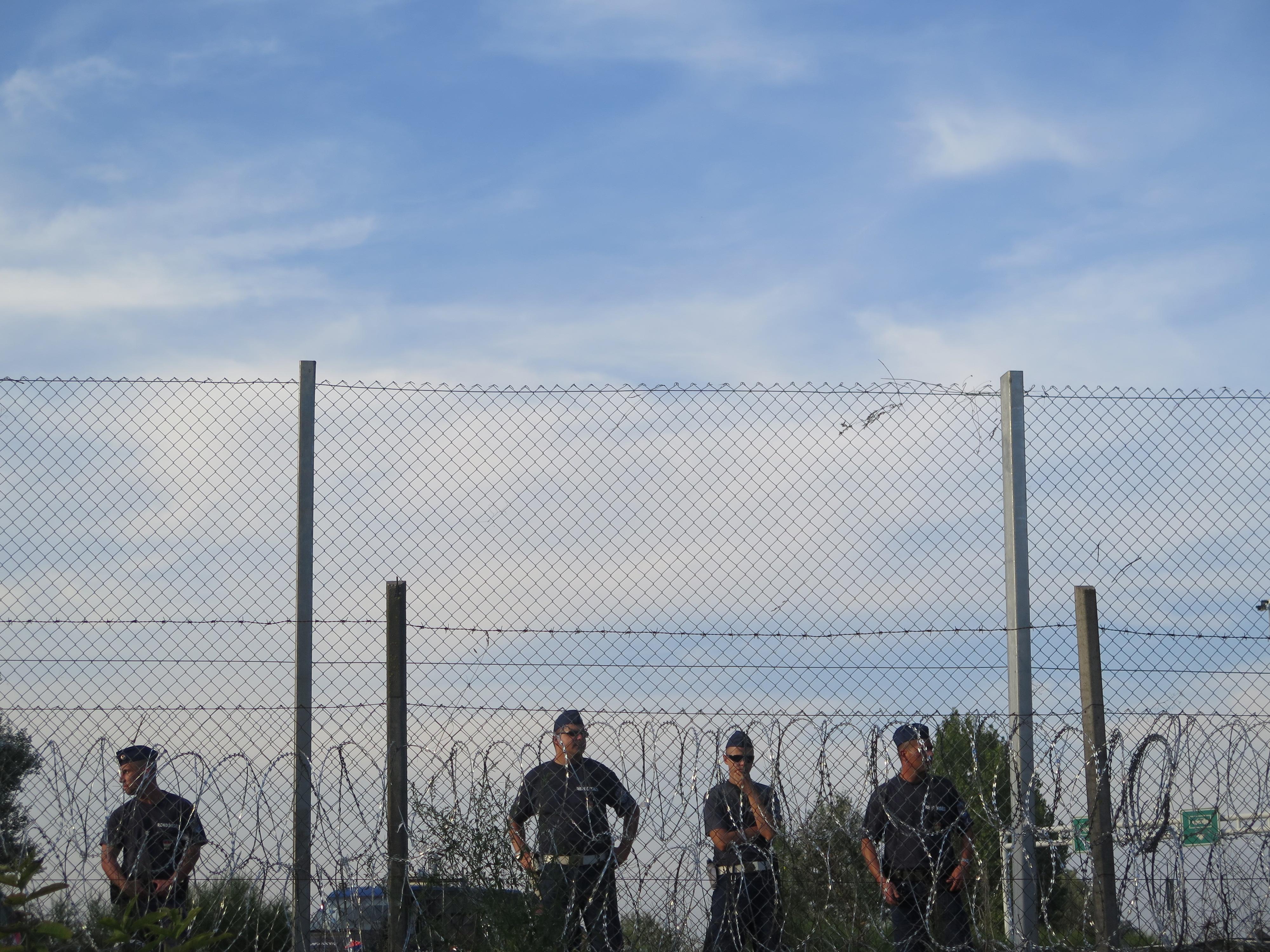 a few border guards stand in line behind multiple rounds of barbed wire fences, over a background with a blue sky. 