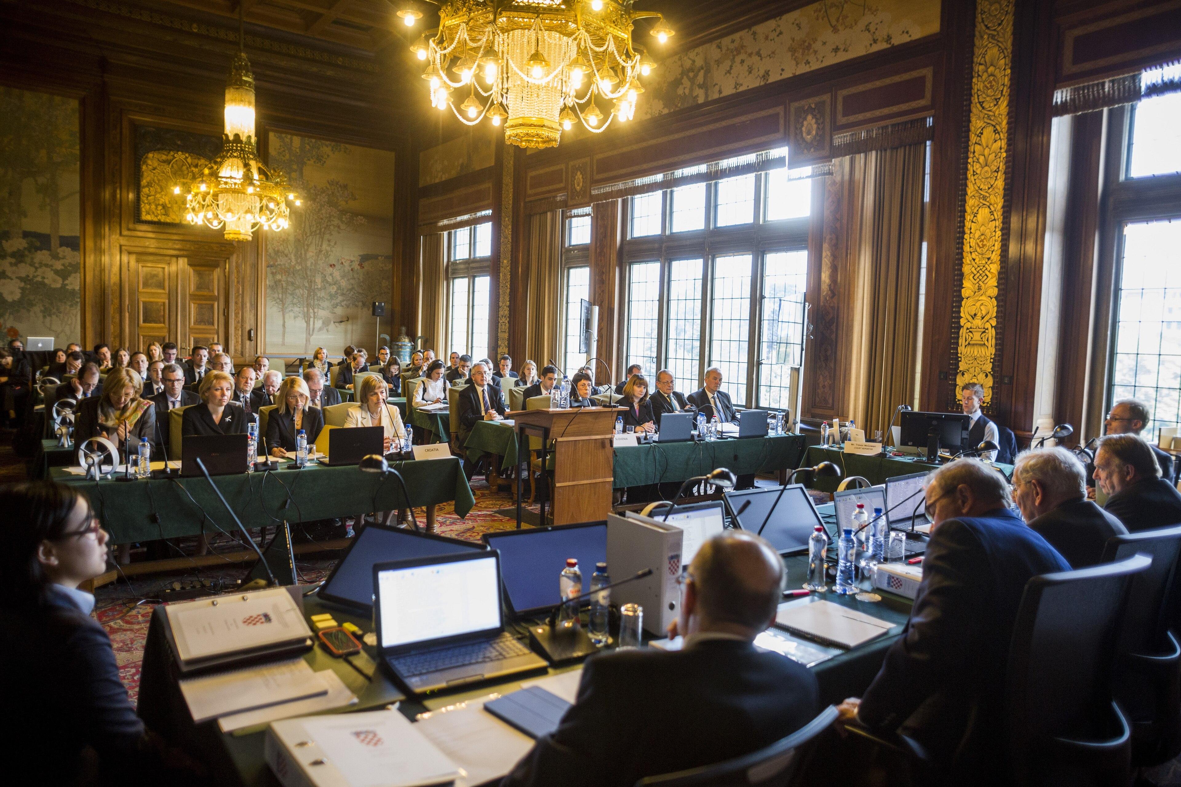 The inside of a court room with chandeliers and high ceilings. Several people sit with laptops and microphones at a table at the front of the room, facing rows of people in the main area of the room