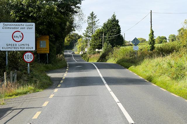 A road undulates over gentle bumps in the landscape. Green trees on the left and green fields on the right. On the left is a circular 100 km/hr sign, and above this, a much larger sign in English and Irish reading: speed limits kilometres per hour