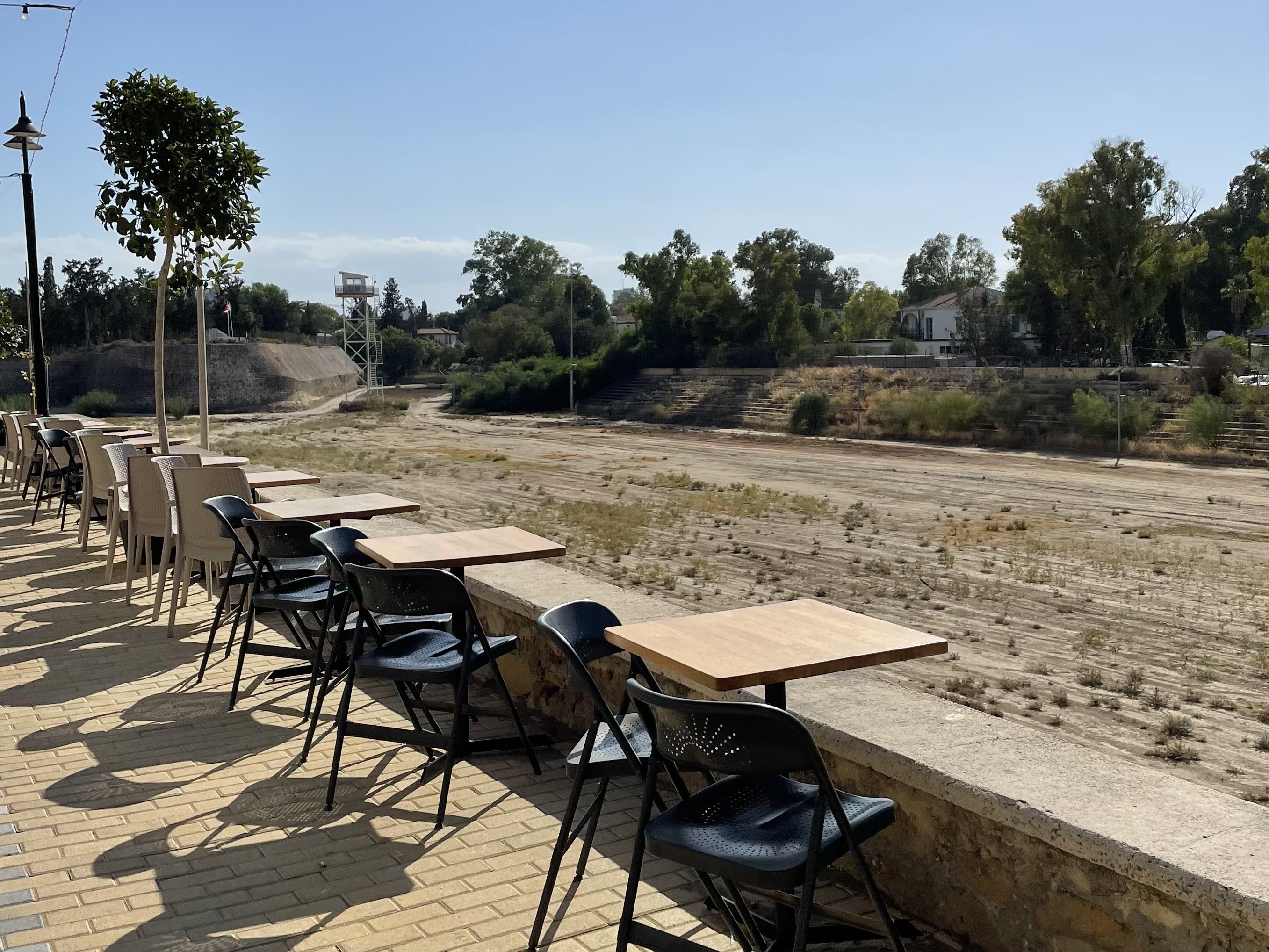 Tables and chairs are lined up along the edge of a brick paved street next to a wall. They overlook a dusty empty patch of land, with scrubby trees, barely distinguishable fence and watchtower in the background
