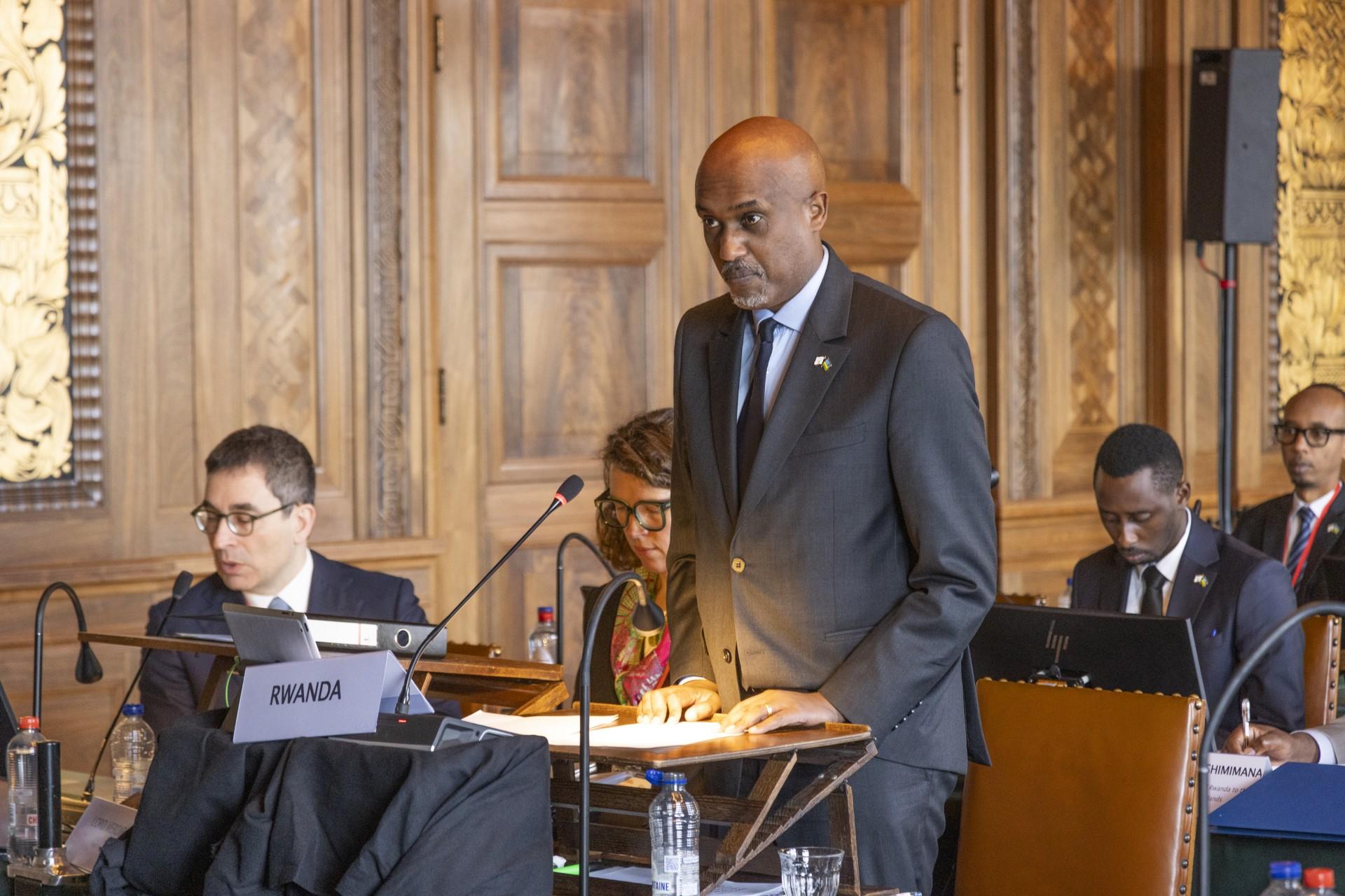 A man is speaking during a hearing in a court, in front of him a sign that says 'Rwanda'