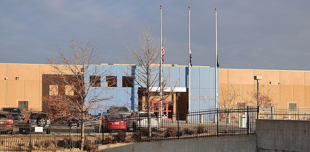 A large building with a US flag flying outside. Lettering on the outside reads: Aurora ICE Processing Center