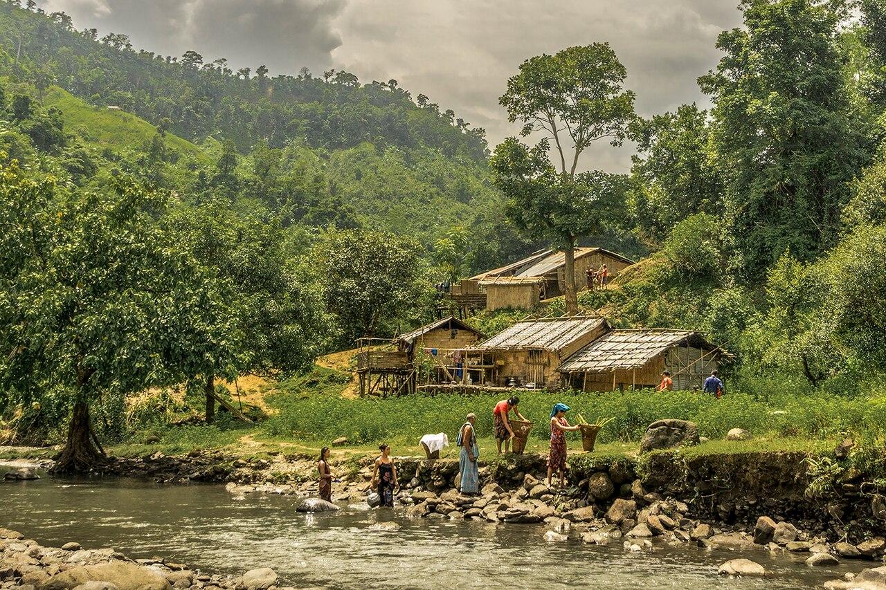 A lush green valley and river next to which people carry baskets. Small huts sit nearby at the foot of the hill
