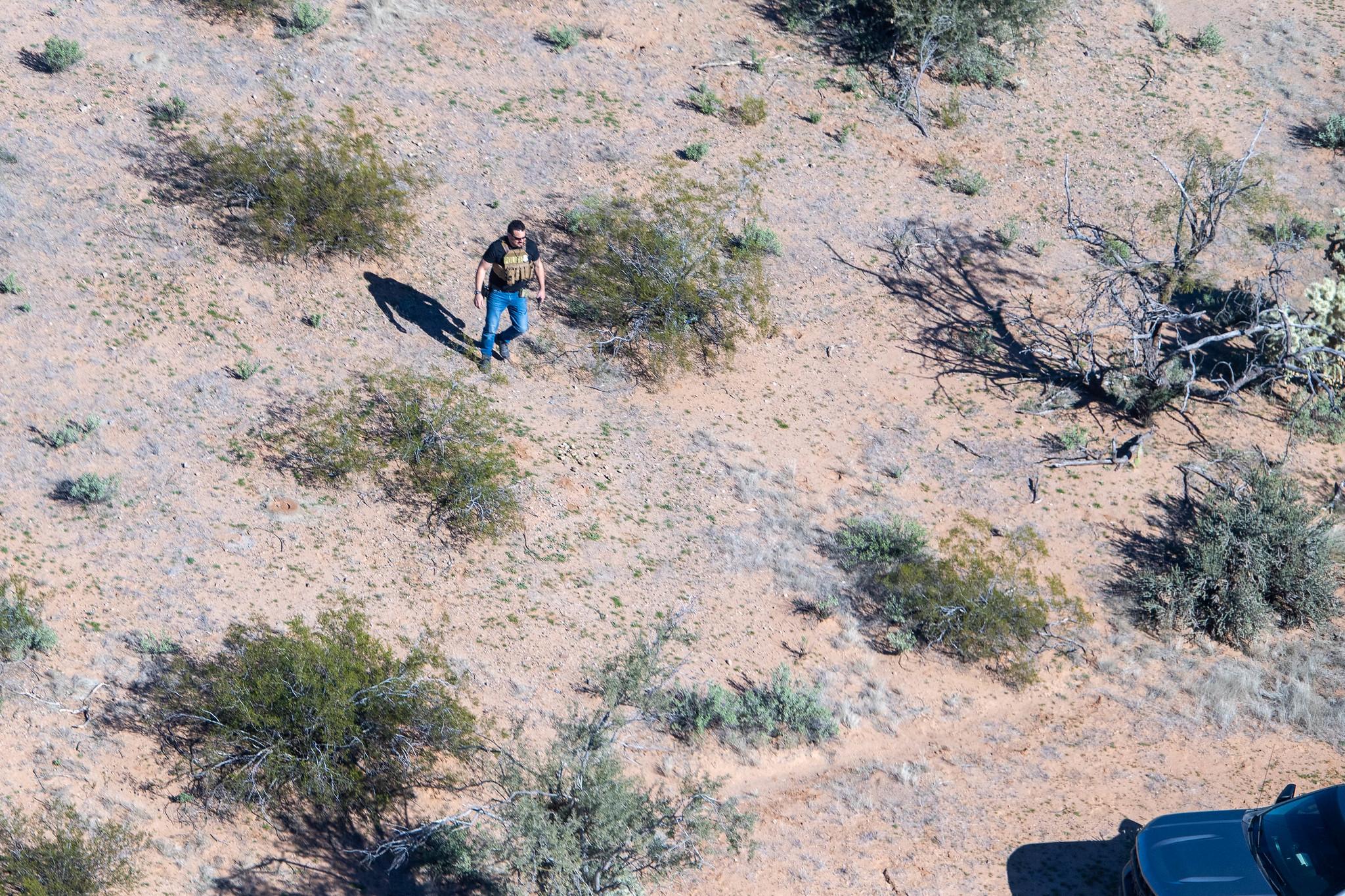 A man wearing sunglasses, jeans, black t-shirt and a beige police vest walks alone in a desert area –&nbsp;brown earth and some green scrub 