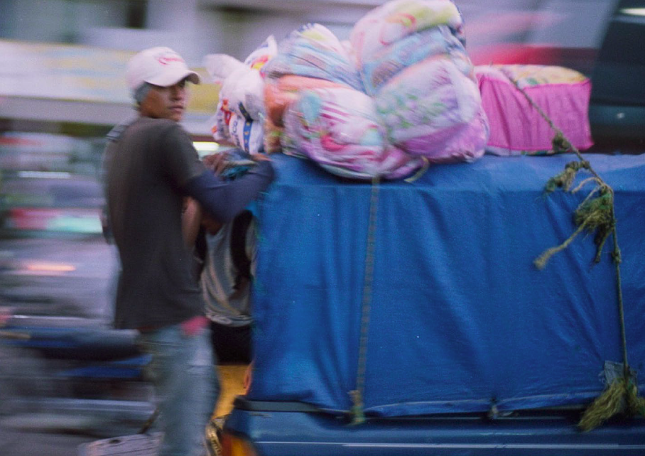 a person wearing a blue shirt and a white cap is holding on to the back of a truck, filled with things and secured with ropes that hold bags on top of it. The truck appears to be moving 