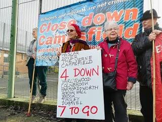 Several people stand in front of a fence holding a big banner reading 'Close down Campsfield'. Two people with a placard stand in front of the banner with a placard reading '4 down. 7 to go' and a list of the detention centres