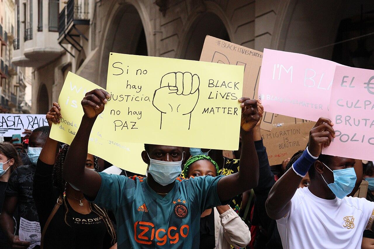 People wearing masks hold cardboard signs reading Black Lives Matter at a gathering in a city. One sign also reads: 'Si no hay justicia no hay paz'