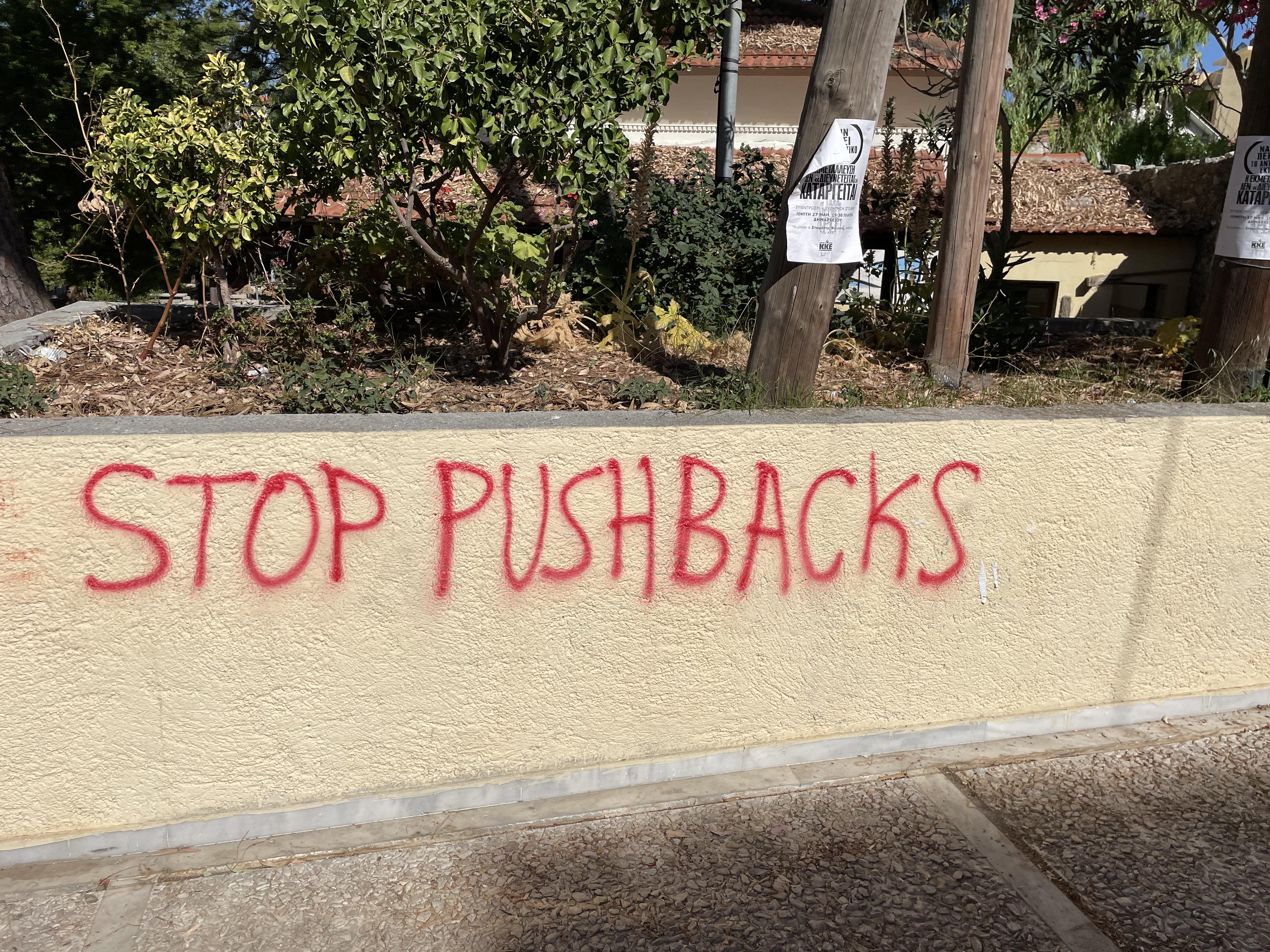 Red graffiti on a low, beige coloured wall reading 'stop pushbacks'. Above the wall are trees and a paper poster in Greek