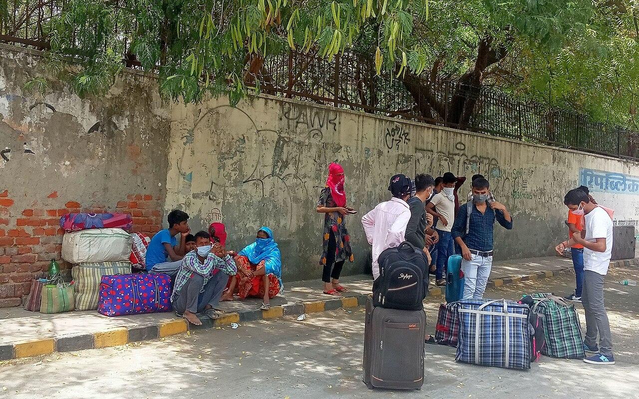 People wearing covid masks stand around with large suitcases next to a worn out bus. Some sit on the pavement next to a graffitied wall 