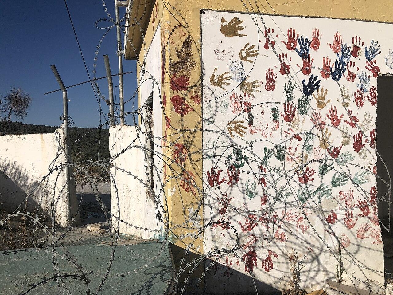 A wall covered in painted hand prints, behind barbed wire