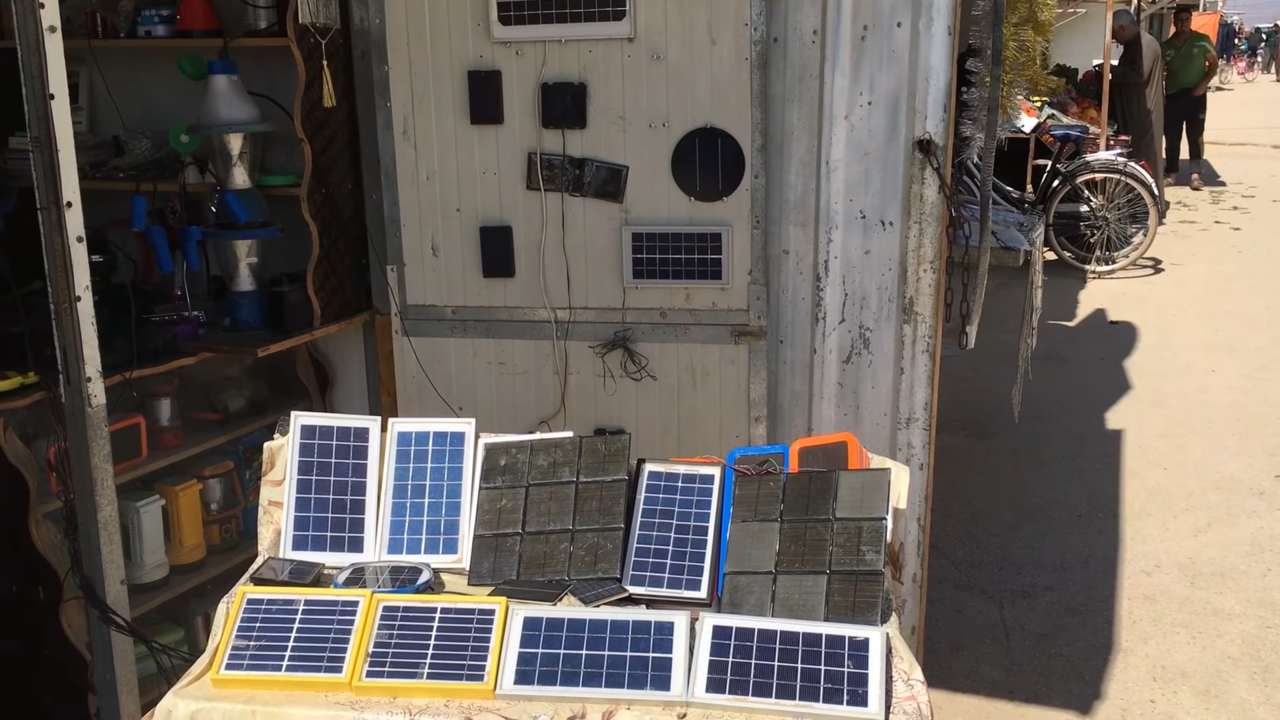 Small solar chargers laid out on a table under a shelter next to a dusty pathway in a refugee camp