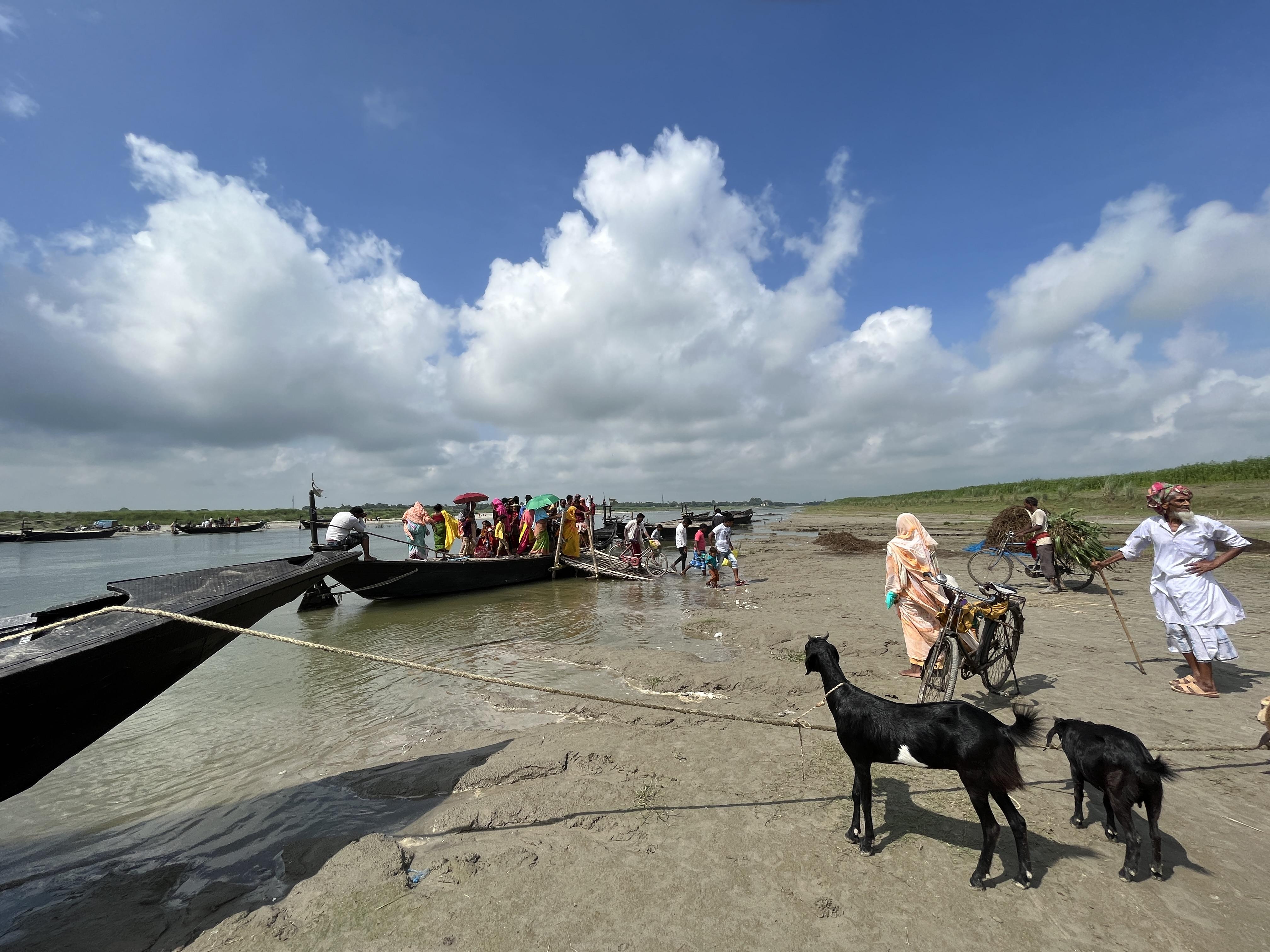 People get on and off a narrow boat moored at the edge of a river, against clouds, blue sky and green banks. A man holding a staff waits. Two goats are tethered to another mooring line. A bicycle waits for its owner