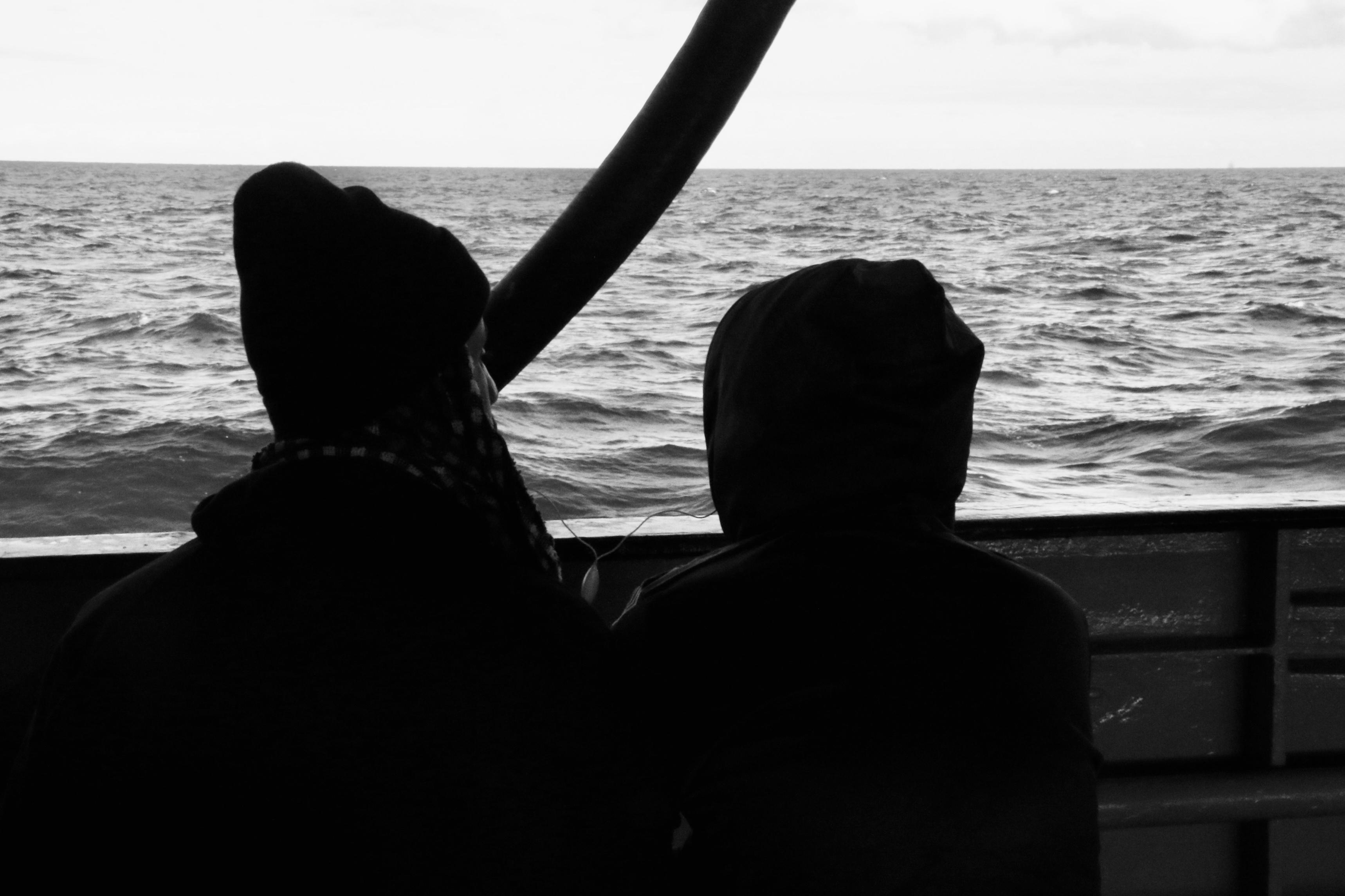 Two people look out to sea from on board a boat in a grayscale scene. They share a wire set of headphones, which hang between their figures against the backdrop of waves