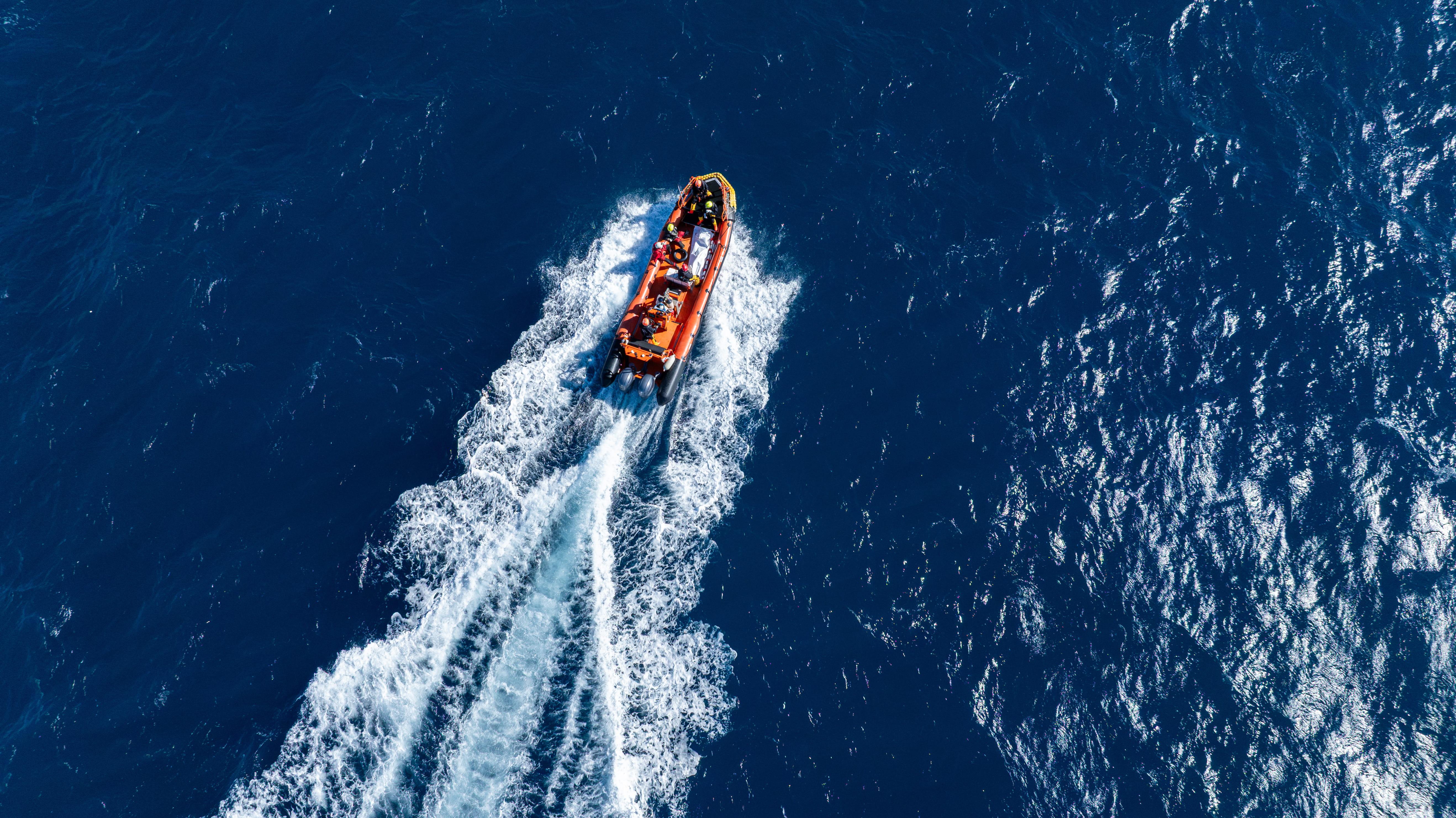 An orange lifeboat speeds through the ocean, as seen from above. At a distance, a white body bag can be seen on board