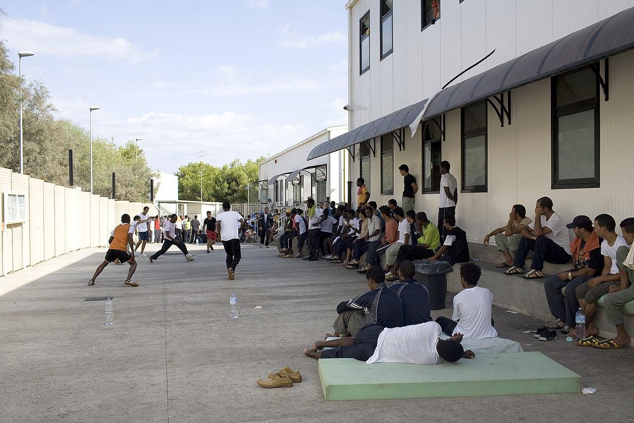 Men sit on mats and on concrete benches around the edge of a small concrete yard while others kick a ball around. Colours are grey and pale white, institutional. Except for the green of the mat and orange of the vest of a player