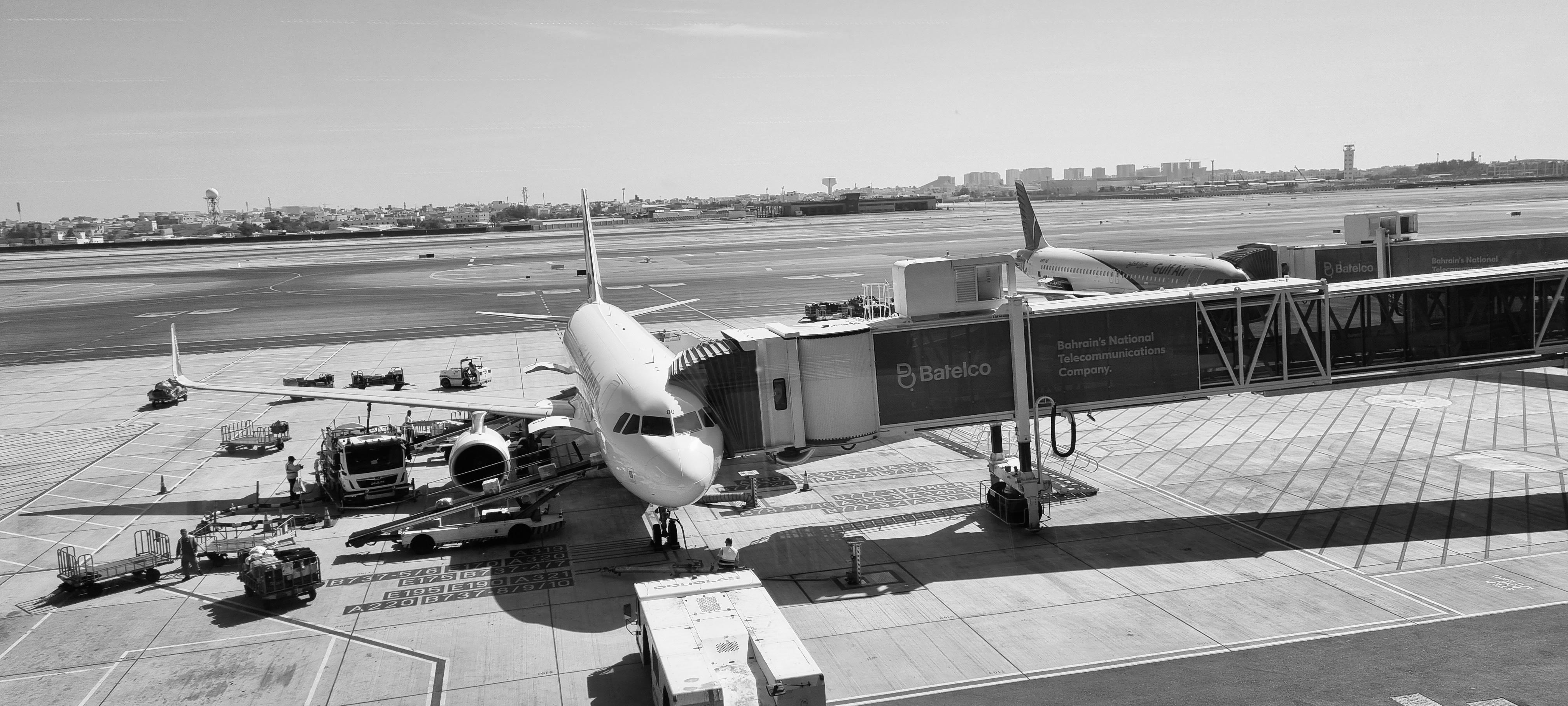 a black and white image of a plane parked at a big airport with a boarding tunnel attached, as seen through a window from the airport building 