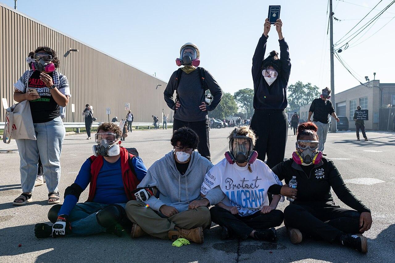 Protestors wearing gas masks sit on the ground with linked arms outside a big warehouse-facility. One person wears a t-shirt reading 'protect immigrants' and another, standing, holds up a US passport