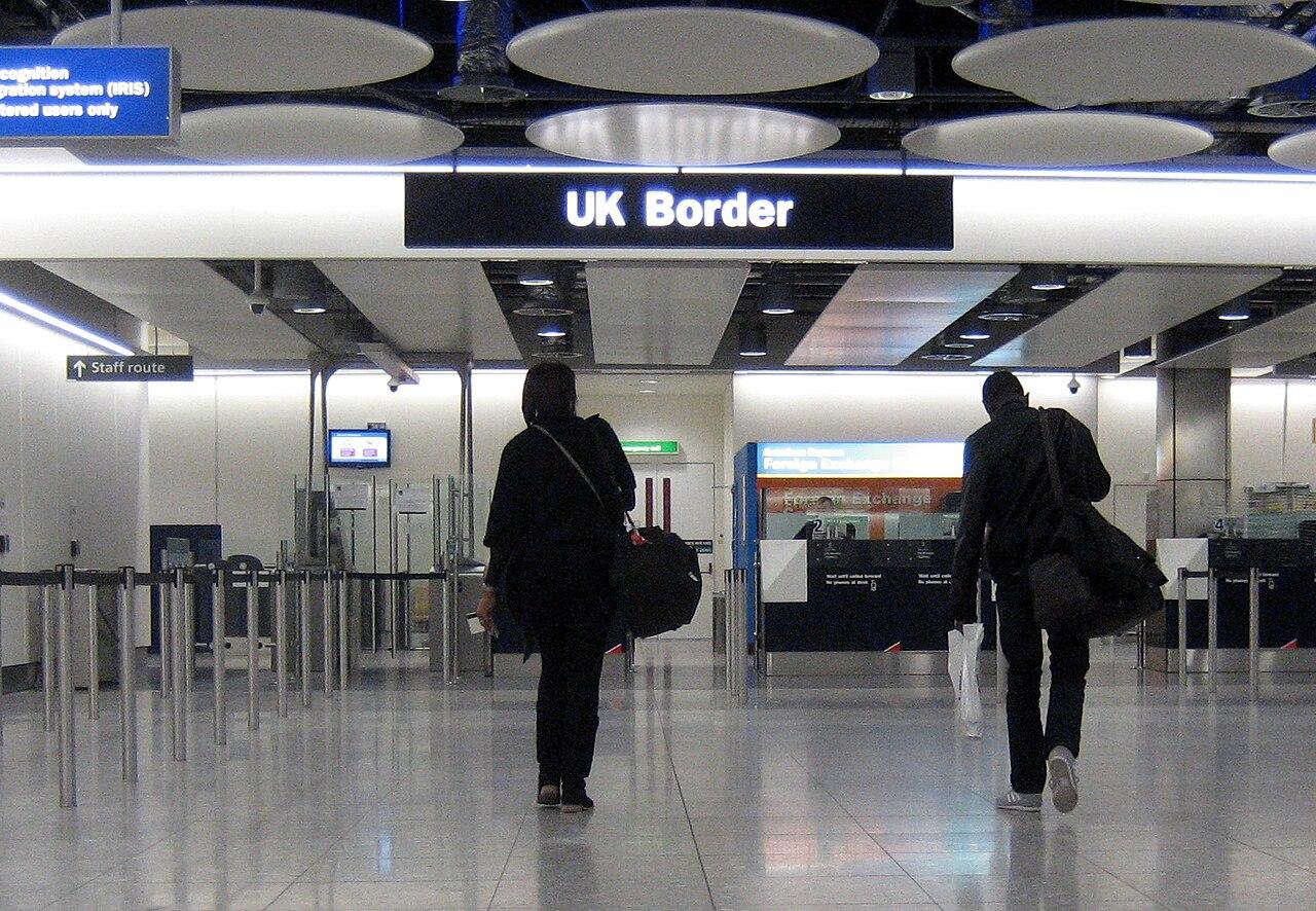 People walk through a brightly lit hall with a blue sign reading 'UK border' overhead