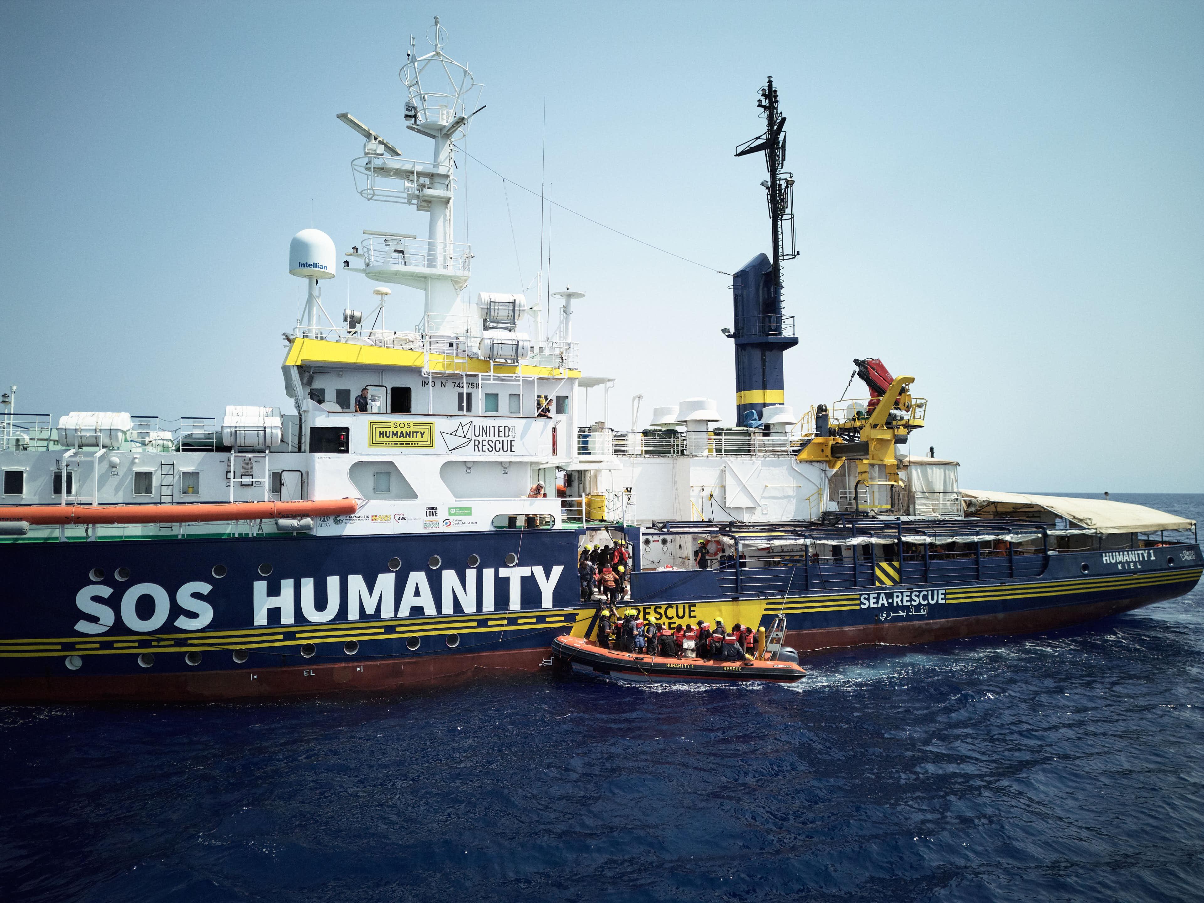 A large vessel, with SOS Humanity painted on the side, at sea. Next to the ship is a small rhib/ dinghy from which rescuers are helping people onto the larger boat.