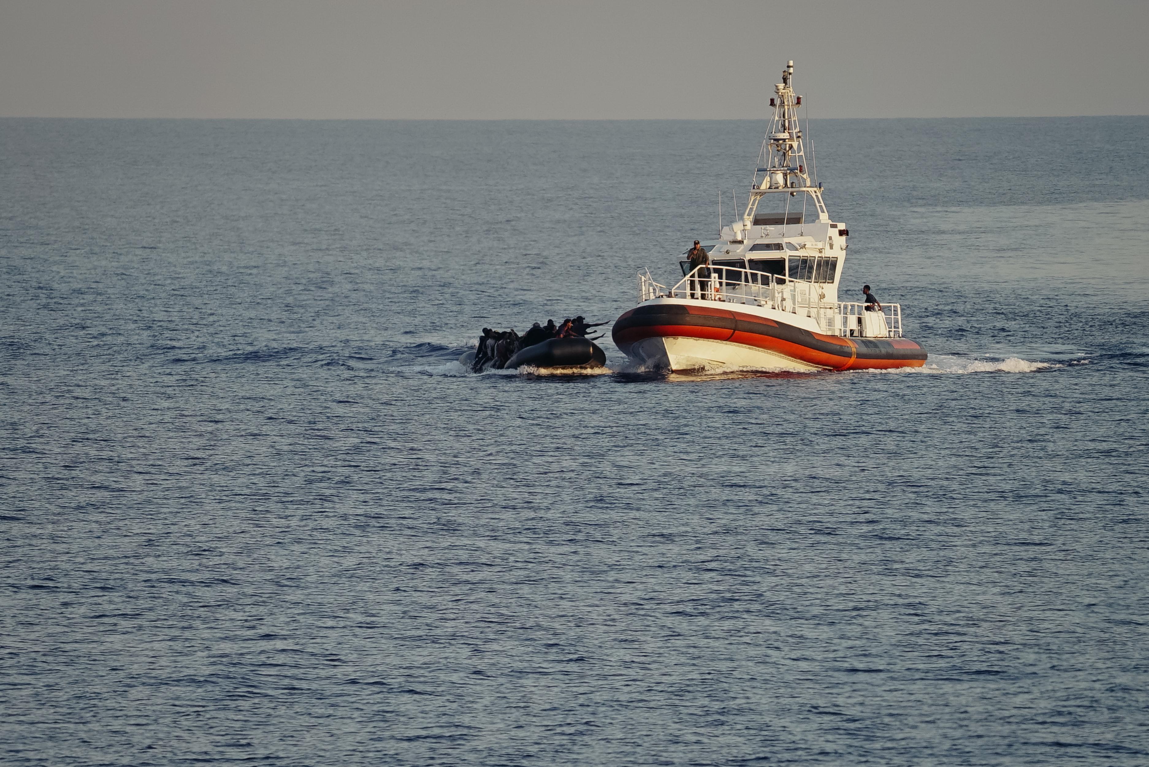 A small overcrowded rubber boat at sea, close to a larger coast guard/ military style boat with two people visible on board. The people on the dinghy stretch their hands out to the edge of the larger boat.