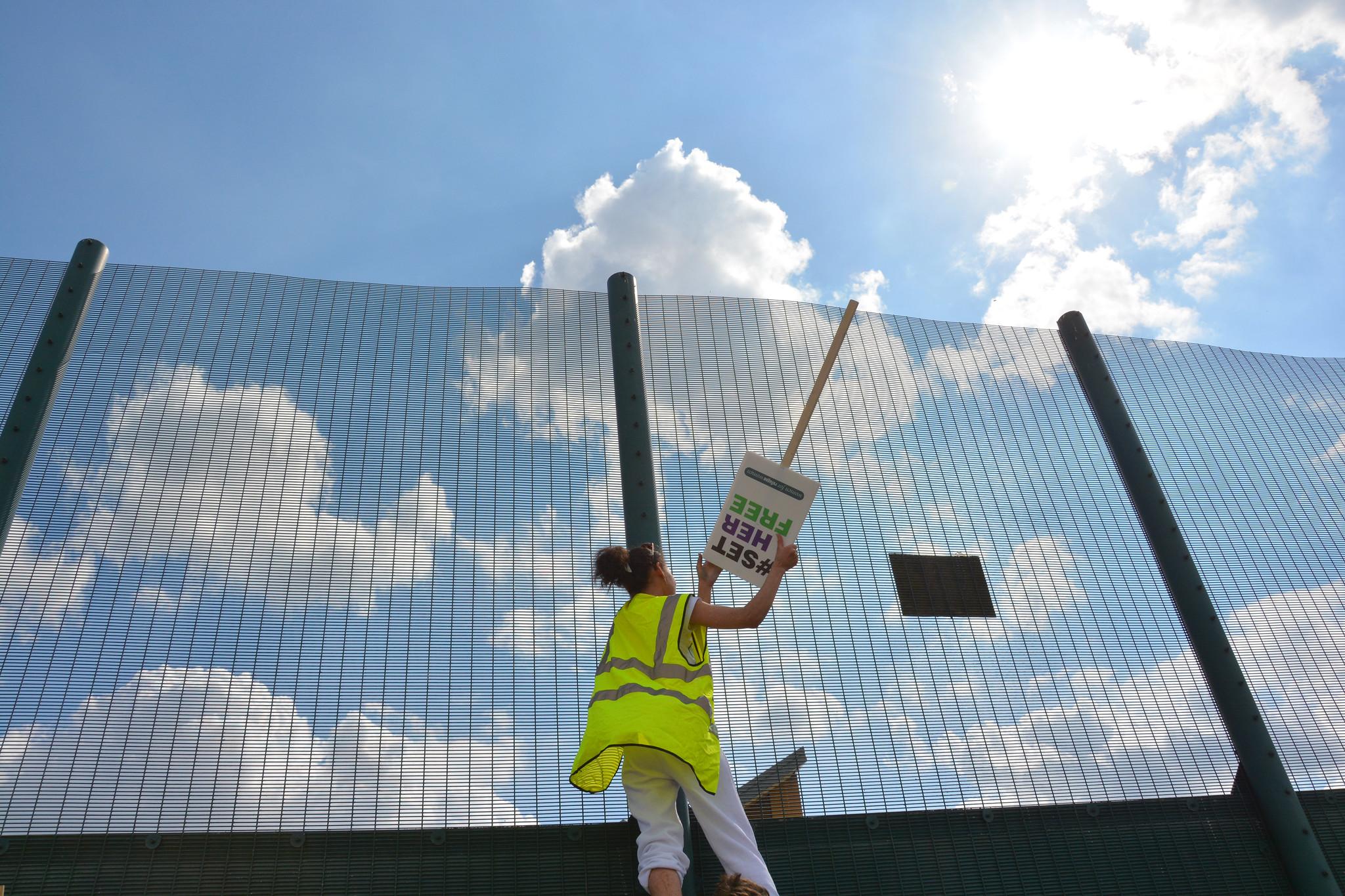 A woman in a high vis jacket is pictured from below standing next to a tall green fence. She holds a sign upside down that reads '#set her free'