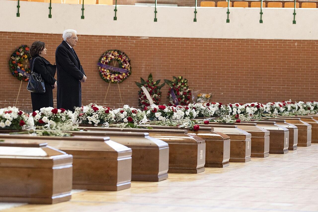 A man and a woman dressed in black stand solemnly by dozens of rows of wooden coffins laid out in a sports hall