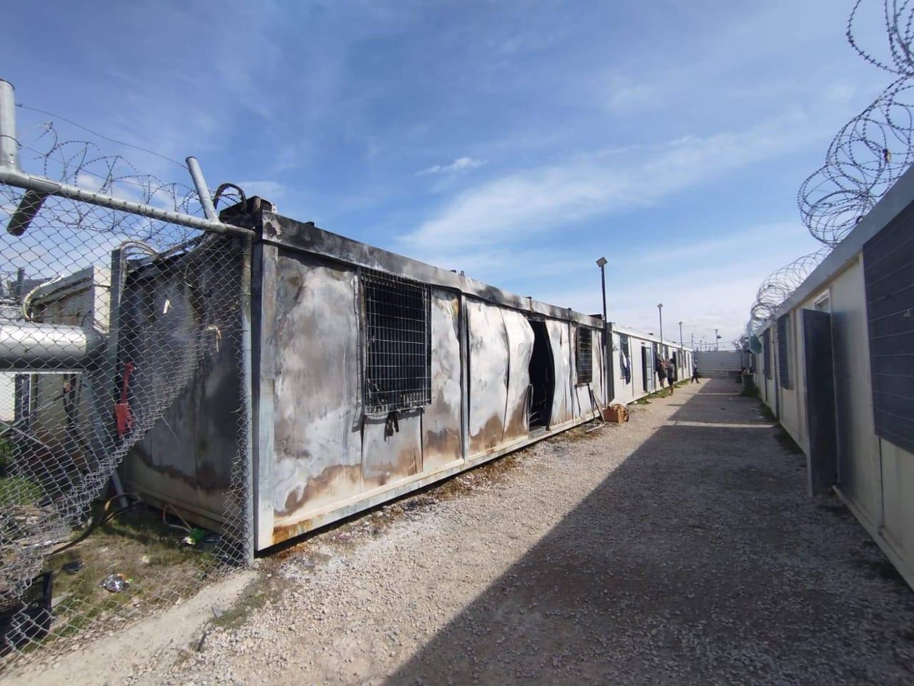 A burnt container is in the centre of the shot with a fence topped with barbed wire to the left. Containers without visible fire damage stand next to it further away, and a figure can be seen going into one