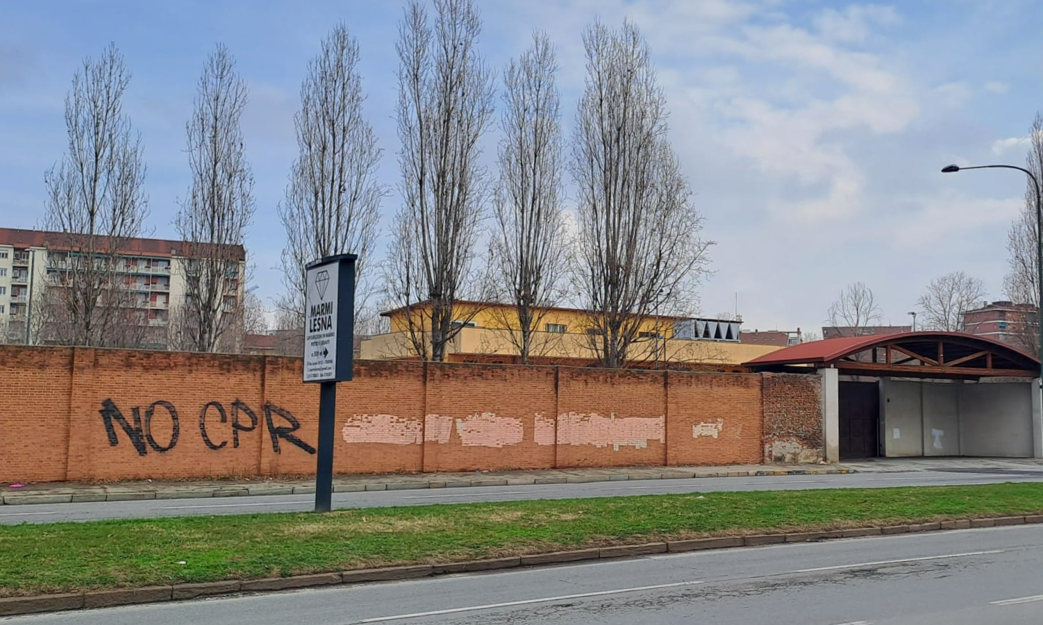 A brick wall with institutional-looking buildings behind it. On the wall is written 'No CPR' (Italian for 'no detention')