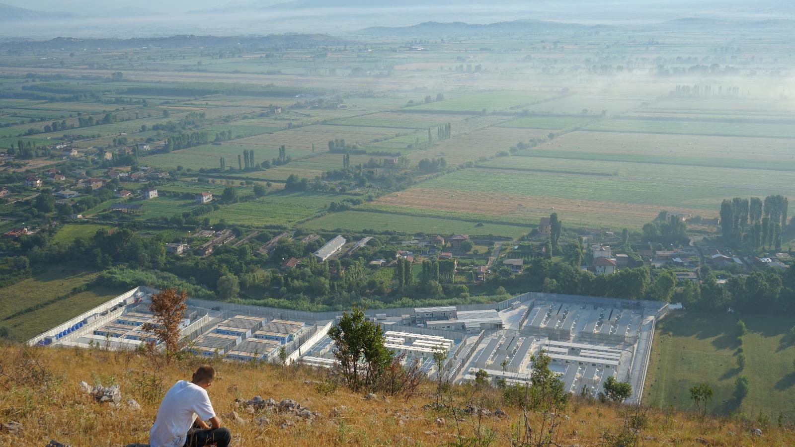 A person sits on a hill overlooking the detention centre in Gjader, Albania: a huge white sprawl of containers and buildings, contrasted against the green of the landscape around.