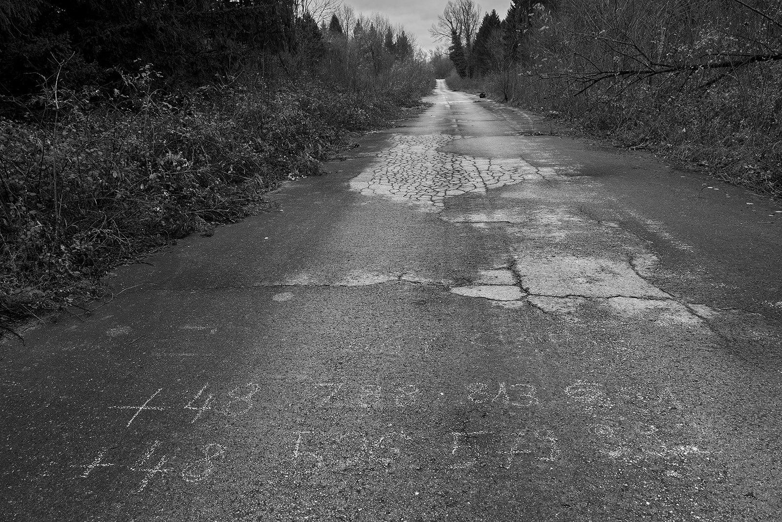 A black and white image of a road at the border of Croatia and Bosnia-Herzegovina. Two phone numbers are written on the road in white chalk –&nbsp;contact numbers for the Balkan route