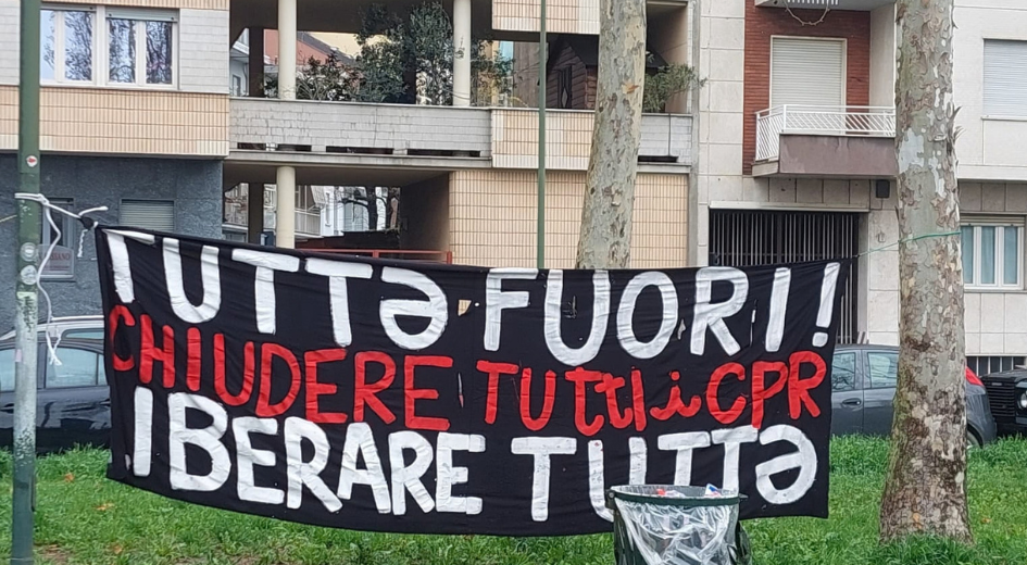 A banner strung between two trees, in front of a block of flats reads in Italian: "Tutta fuori! Chiudere Tutti i CPR. Iberare Tutta" / “Everyone out. Close all CPRs. Free them all”