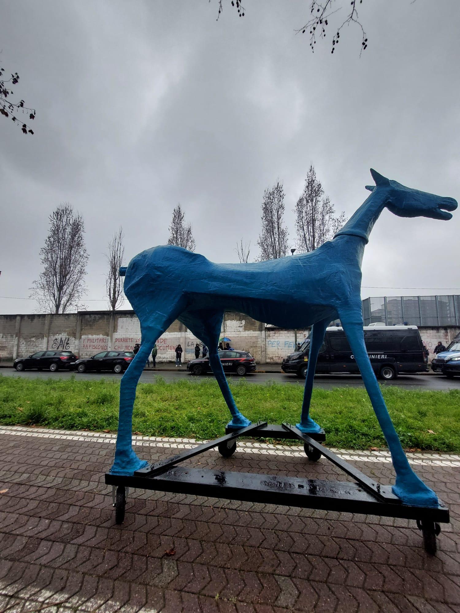 A metal statue of a blue horse stands in front of grass, parked cars and high barbed-wire topped detention centre walls with graffiti painted on them
