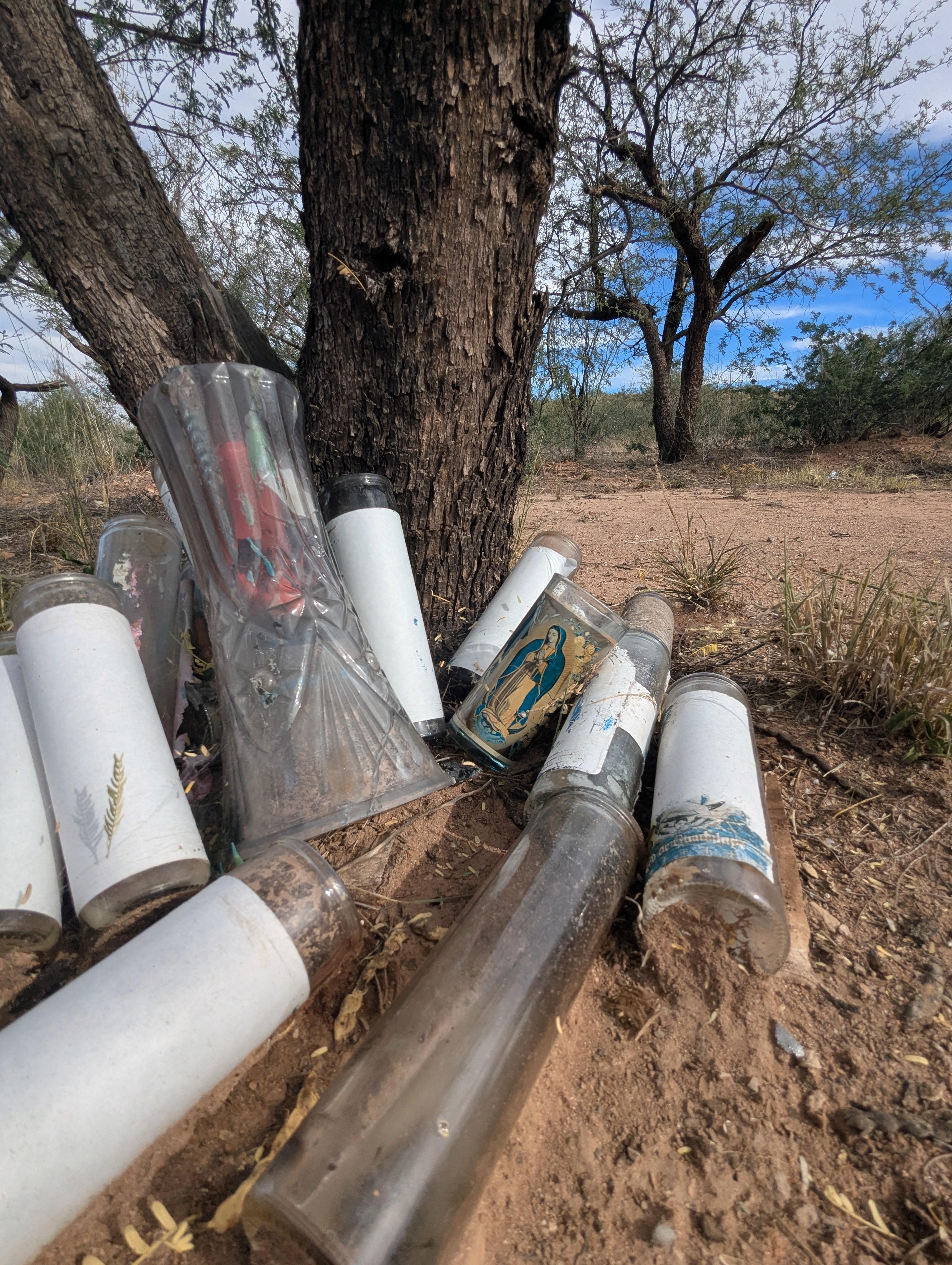 Empty glass containers, some holding the remains of candles and some bearing the image of the Virgin Mary, are piled at the base of a tree trunk