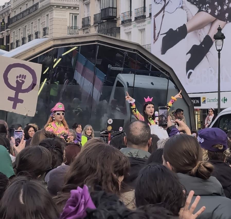 People stand in a crowd watching a woman wearing bright colours stood on a stage or podium, next to others wearing bright colours. Someone holds up a placard with the female sign painted on and a fist inside