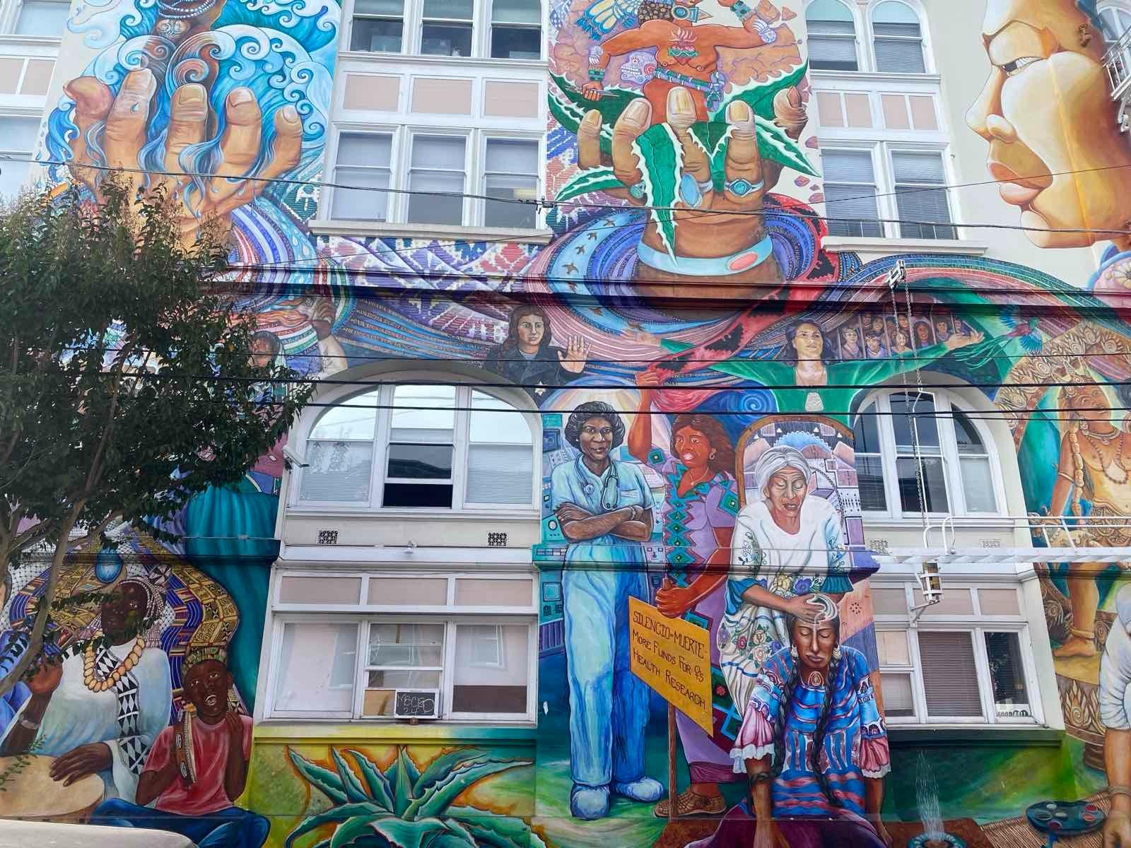 A colourful mural on the side of a building shows women in different poses –&nbsp;one wearing scrubs and a stethoscope next to a woman in a colourful dress holding a placard. One woman appears to be massaging another woman's head, and above them are large hands holding plants and a goddess figure in one, and water and a goddess figure in another