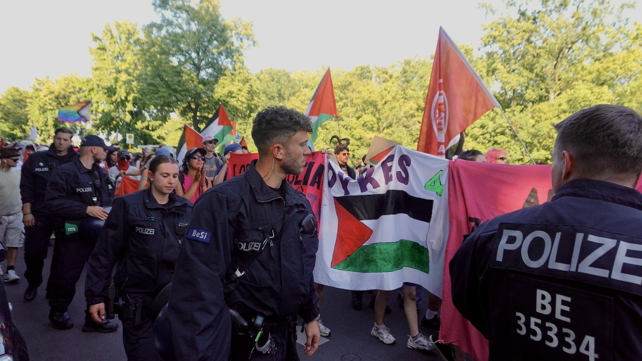Protesters at Berlin's queer pride march walk along a street behind a big banner with the word 'Dyke' and a Palestine flag visible. In the foreground, a line of police officers walks alongside them 